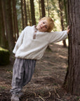 Child in a white sweater and gray pants standing next to a tree in a forest