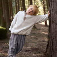 Child in a white sweater and gray pants standing next to a tree in a forest