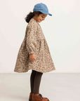 A young child wearing a beige and brown leopard-print dress, a light blue cap, dark brown tights, and brown leather shoes, standing in profile against a plain white studio background.