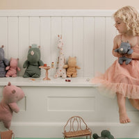 Child in a pink dress sitting on a white shelf with stuffed animals in a room with green floor and beige walls.
