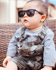 Baby wearing sunglasses and overalls sitting on a wicker chair.
