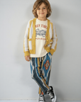 A young boy stands against a light gray wall wearing a cream crewneck sweatshirt with a tiger graphic and 'ASY TIGRE' text, layered under a mustard yellow and white fuzzy cardigan. He is also wearing patterned blue, yellow, and gray pants with black and white sneakers.