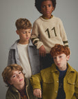 Four boys modeling knit and casual outerwear in a studio setting against a solid gray background. The group includes a boy in a cream sweater with the number 11, a boy in a gray jacket over a white shirt, a boy in a green corduroy jacket, and a boy in a mustard green corduroy jacket over a dark shirt.
