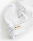 A close-up of a white knit baby hat with a soft blue whale print and a beige brand tag reading MartMar.
