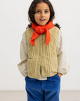 A young girl with dark curly hair stands against a plain white studio background, wearing a cream-colored textured knit cardigan vest over a white long-sleeve shirt, a bright orange knitted scarf tied in a bow, and blue corduroy pants.