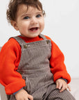 Child wearing a red shirt and brown striped overalls sitting on a white surface.