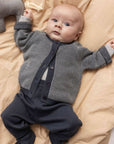 A baby lying on beige bedding, wearing a gray textured knit cardigan over a white undershirt and dark navy pants.