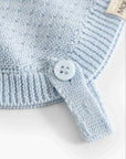 Close-up of a light blue knit baby garment with a ribbed cuff, a matching light blue button tab, and part of a beige fabric tag.
