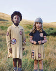 Two children standing in an outdoor grassy field wearing matching beige patchwork dresses and a navy top with striped sleeves, both featuring circular embroidered patches.