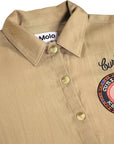 Close-up of a beige button-up shirt with a collar, showing the Molo brand label inside the neck, decorative script embroidery on the chest, and a circular patch with a smiley face.