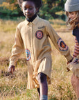 Two children outdoors in a grassy field, wearing matching tan scout-style uniforms with circular embroidered patches. One child is standing and the other is crouching, both looking down at the ground.