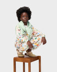 Child wearing a floral outfit with a green scarf, sitting on a wooden stool against a white background