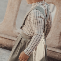 Child wearing a plaid shirt and gray dress standing in front of a stone wall.