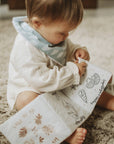 A baby sitting on a textured beige rug, holding a white cloth book with black illustrations and brown botanical designs. The baby is wearing a white long-sleeve bodysuit and a light blue patterned bib.
