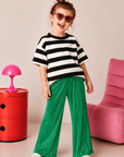 Child wearing a black and white striped shirt and green pants standing in a room with a red side table and pink chair.