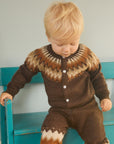 Child wearing a brown patterned outfit standing on a wooden floor with a teal bench.
