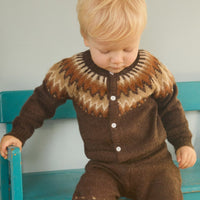 Child wearing a brown patterned outfit standing on a wooden floor with a teal bench.