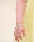 A close-up of a child's hand and wrist wearing a delicate silver-toned bracelet adorned with small white and yellow floral details, set against a soft cream background and a pale yellow dress.