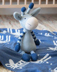 A crocheted giraffe plush toy in light gray and navy blue sits on a blue blanket with white giraffe patterns, placed near a crib.