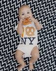 A baby lying on a black and white chevron blanket, wearing a white bodysuit with a pretzel and 'NY' graphic, while holding a pretzel toy.