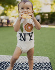 A baby standing outdoors on a black and white chevron blanket, wearing a cream bodysuit with a New York City skyline graphic and 'NY' text, while holding a bagel to their mouth.