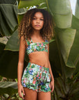 Young girl in a floral two-piece outfit standing among large green leaves.