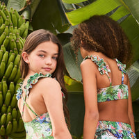 Two children wearing floral swimsuits standing in front of banana trees and large leaves.