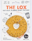 A packaged baby teether shaped like a donut, with a mustard-yellow surface, white sesame-seed pattern, and a small wooden stick handle. The item is mounted on a white printed backing card with orange and black text reading 'THE LOX' and 'NATURAL RUBBER BABY TEETHER,' plus illustrated doodles and a 'Peculiarity' logo.