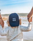 Child wearing a 'BIG BRO' cap on a beach with two adults holding hands