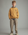 A young boy stands against a neutral gray studio background wearing a mustard yellow sweatshirt with a navy blue collar, beige and navy vertically striped trousers, and beige sneakers.