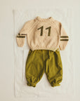 A baby knit two-piece outfit featuring a cream-colored long-sleeve sweater with the number '11' and dark olive stripes on the sleeves, paired with olive green pants, laid flat on a white sheet.