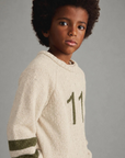 A child model wearing a cream-colored knit sweater with olive green stripes on the sleeves and a large olive green number '11' on the chest, set against a plain gray studio background.
