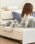 Child sitting on a bed with 'Avocado' branding, wearing a white sweater and gray leggings.