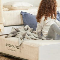 Child sitting on a bed with 'Avocado' branding, wearing a white sweater and gray leggings.