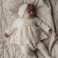 Baby in a white outfit with a textured skirt lying on a beige blanket.