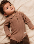 Baby wearing a brown ribbed outfit with a collar, lying on a soft surface.