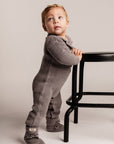 Child wearing a gray ribbed outfit standing next to a black chair on a plain background