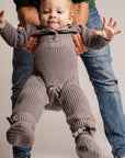 Baby in a knitted outfit being held up by an adult on a plain background
