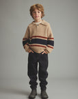 A young boy stands against a neutral gray studio background wearing a beige ribbed knit sweater with horizontal navy and rust-red stripes, dark pants, and dark boots.