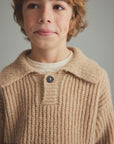A close-up of a child wearing a beige ribbed knit polo cardigan with a dark button, layered over a cream shirt, against a neutral gray background.