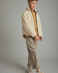 A child model wearing a cream sherpa fleece zip-up jacket over a mustard yellow shirt, paired with beige and brown vertically striped trousers and beige sneakers, set against a plain gray studio background.