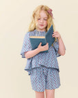 A young girl with blonde hair and a red-and-white bow stands against a plain beige background, holding a blue book. She is wearing a light blue patterned short-sleeve top and matching shorts.