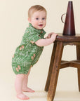 A baby standing on a wooden stool and holding onto it, wearing a green short-sleeve romper with a white musical notation print. A dark brown conical object sits on the stool.