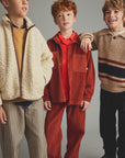 Three boys modeling children's clothing against a plain gray studio background. The boy on the left wears a cream sherpa zip jacket over a mustard top and striped pants.