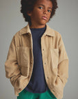 Child wearing a beige corduroy jacket over a navy shirt with green pants on a gray background