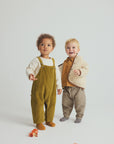 Two toddlers modeling coordinated autumn outfits in a studio setting. The child on the left wears olive corduroy overalls over a cream shirt, and the child on the right wears a cream textured cardigan over a mustard top with brown-and-beige striped pants.