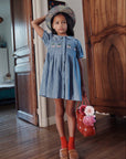 Young girl in a denim dress standing in a room with wooden furniture.