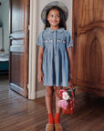 Young girl in a blue dress standing in a room with wooden floors and doors.