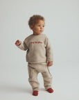 A toddler standing against a light gray studio background, wearing a beige knit two-piece outfit with red text reading 'joy in play' on the chest and red socks.