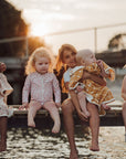 Woman holding a baby and sitting with two other children by a body of water at sunset.